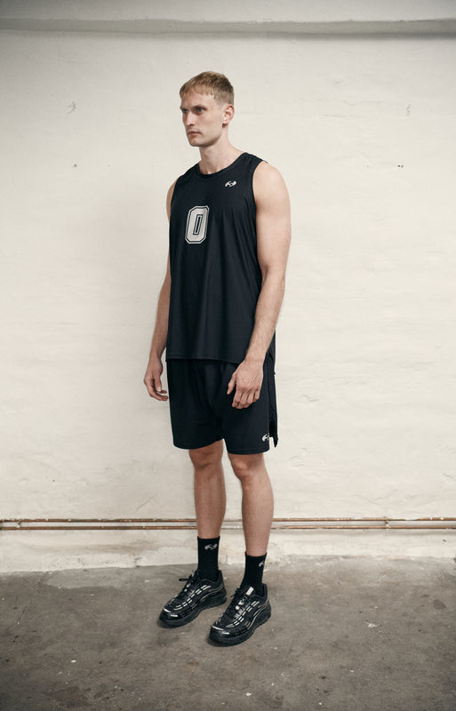 Man wearing a black sleeveless shirt and shorts with logos, standing against a white wall.