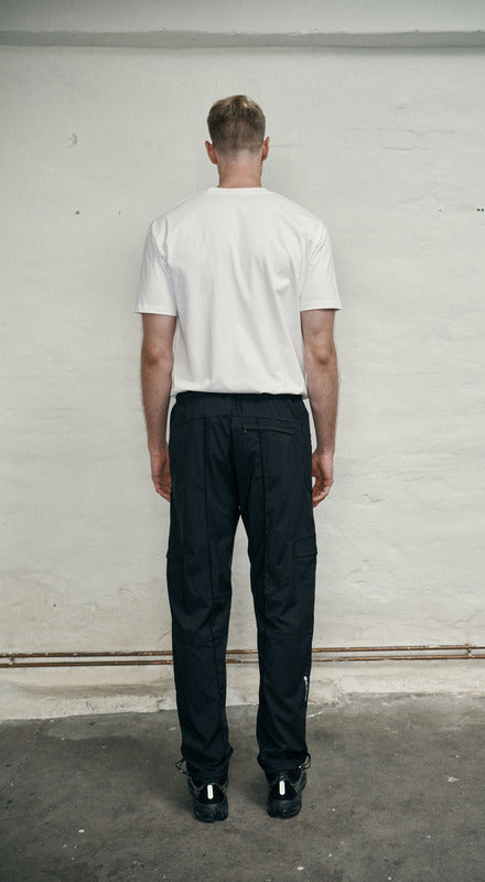 Person wearing a white t-shirt and black pants standing against a plain wall.